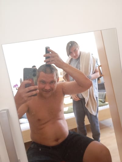 In a room, a man in his 40s is shaving his head in front of a mirror, holding a phone in his left hand and a shaver in his right. He is shirtless and wearing black shorts. In the reflection of the mirror, another person, who appears to be a younger individual, is standing behind him, wearing a grey t-shirt and dark pants, with a towel draped over their shoulders. The room appears to be a bedroom, with a bed and a wooden floor visible in the reflection. The image is taken in Bitterfeld-Wolfen, Germany.