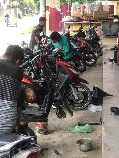 The image depicts an outdoor motorcycle repair shop in Rambot, Indonesia. There are multiple motorcycles in various states of repair, parked close together. Three men are actively working on the motorcycles. One man wears a green shirt and appears to be examining the engine of a red and black motorcycle. Another man is wearing a black uniform shirt and appears to be assisting in some way. The third man, wearing a striped shirt, is working on the front of a motorcycle. 

Various tools and motorcycle parts are scattered on the concrete floor around the bikes. There is a bucket, a wrench, and a piece of plastic on the floor. In the background, there is a metal shelving unit with various motorcycle parts and containers, including a "POLY TOP" box. The front of the building is partially visible, with trees and other buildings in the distance.