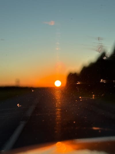 A photograph captures a sunset over a rural landscape, possibly in or around Морозовка, Russia. The sun, a bright white orb, sits just above the horizon, casting a warm orange glow across the sky and reflecting off a body of water or a wet road. The scene is viewed through a window, as evidenced by the streaks and specks of dirt or water droplets on the glass. In the foreground, the edge of a car or vehicle is visible, with a blurry orange reflection. The background features a dark silhouette of trees or hills against the colorful sky. The overall impression is one of a serene and beautiful natural scene, possibly experienced during a drive.