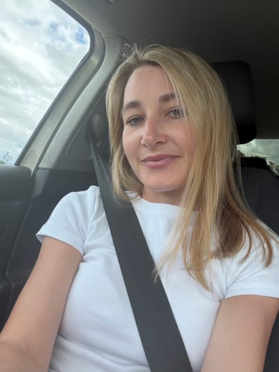 A young woman is depicted in a selfie or portrait taken inside a car, likely in Avdon, Russia. The image is oriented sideways, with her head tilted to the right. She has long, straight blonde hair, light-colored eyes, and visible freckles across her nose and cheeks. A gentle smile plays on her lips, suggesting a calm or content mood. She is wearing a white, ribbed short-sleeved top and has a dark seatbelt securely fastened across her chest. The car interior includes a dark seat and a visible headliner. Through the car window, a cloudy daytime sky is visible. Faint text, including what appears to be a "VW" logo, is discernible on a sticker on the car window in the lower left of the original image.