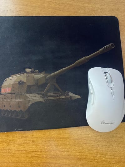 The image shows a white computer mouse placed on top of a black mousepad that features a graphic of a tank. The mouse is positioned in the upper right quadrant of the image, with its scroll wheel and buttons clearly visible. The mousepad displays a detailed image of a military tank, with its turret and cannon prominently featured. The background of the image is a wooden surface, suggesting the mousepad and mouse are on a desk or table. There is no indication of the time of day, weather, or specific location beyond the provided context of Mamontovo, Russia. The image is a still photograph, so no activity or event is taking place. The only visible text is the brand name "smartbuy" printed on the mouse.