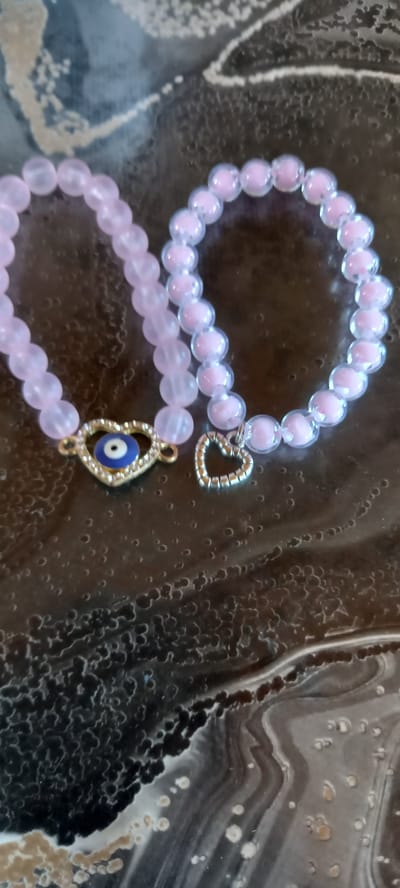 Two pink beaded bracelets are laid out on a dark, patterned surface. One bracelet has a gold heart charm with a blue and white evil eye charm in the center, and the other has a simple silver heart charm. The surface appears to be a tabletop or countertop with a textured, swirling pattern in shades of dark grey, black, and white. The lighting is overhead and casts a slight shadow. There are no people or animals visible in the image. The location of Iskra, Bulgaria is not discernible from the image itself.