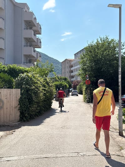 This vertically oriented, eye-level media file depicts an outdoor scene on a sunny day, likely in the afternoon, given the long shadows. The setting appears to be a residential area with multi-story buildings and lush green vegetation. In the foreground, a person wearing a yellow t-shirt with red lettering and red shorts is walking on a paved sidewalk. The t-shirt has text that reads "SPASILAČKI LIFE GUARD". The person is also wearing dark-colored Crocs. Their back is to the camera, and they are walking away from it. Further down the sidewalk, another person is riding a bicycle, also with their back to the camera. This cyclist is wearing a red t-shirt and light-colored shorts. The background features several modern residential buildings with balconies, and a verdant, tree-covered hillside rises in the distance under a clear blue sky with a few wispy clouds. The overall atmosphere is casual and relaxed, suggesting a warm, pleasant day in a Mediterranean or similar climate. The visible text on the t-shirt indicates a role related to lifeguarding.