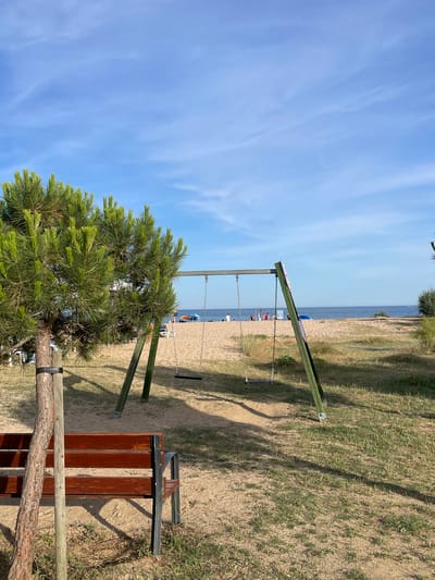 This is a photograph of a swing set on a sandy beach with a pine tree in the foreground. The swing set is green and has two swings. The beach is populated with a few people and umbrellas. The ocean is visible in the background. The photograph appears to have been taken on a sunny day. The scene is located in Arenys de Mar, Spain.