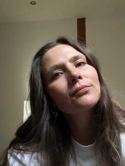 The original image, rotated 90 degrees clockwise, features a close-up portrait of a young woman. Her face, framed by long, dark brown hair, is positioned centrally and looks upwards towards the viewer with a thoughtful or slightly curious expression. She has brown eyes and light, naturally colored lips with a subtle pink tint. Her skin has a natural sheen, suggesting good lighting.
She is wearing a white top, visible on her right shoulder and upper chest area. The background suggests an indoor setting with light-colored walls. To her left (when the image is conceptually rotated upright), a dark brown, rectangular wooden beam is visible running horizontally across the wall, indicating rustic or traditional architecture. On the upper left wall, a small, circular, dark-colored object, possibly a light fixture or vent, can be seen. The lighting appears bright and natural, suggesting it is daytime. No visible text is present in the image. The setting could be a room in Bartula, Montenegro, given the location context.