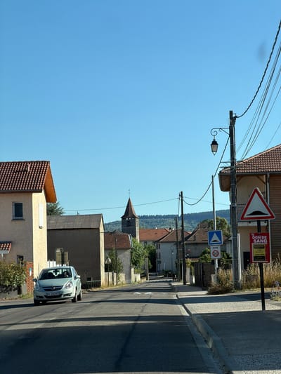 This image shows a street scene in a French village. The perspective is tilted, making it appear as though the viewer is looking down from a slight angle. The sky is a clear blue, indicating a sunny day, likely in the afternoon due to the long shadows cast across the road.

The foreground features a light-colored car parked on the right side of a paved road. The car has its headlights on and its license plate is visible, reading "HC-282-WT". A concrete curb runs along the edge of the road.

The middle ground shows several buildings with red tile roofs, typical of rural French architecture. A distinctive church tower with a pointed roof rises above the other structures. There are also street signs visible. One sign, in a triangular red border, depicts a horn. Another sign is blue with a white triangle and the word "ZONE" above a "30" indicating a 30 km/h speed limit. A red sign with white text announces "DON DE SANG" (blood donation) with dates and times.

The background is dominated by a lush green forest covering rolling hills, suggesting the village is nestled in a natural landscape. Telephone wires crisscross the sky, supported by utility poles. The overall impression is one of a quiet, sunny day in a rural French community.