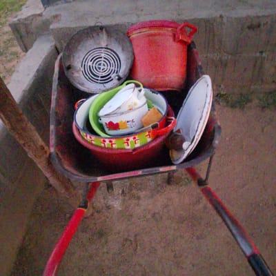 A wheelbarrow is loaded with household items, including a red bucket, a metal sieve, and several bowls and pots. The wheelbarrow is positioned on a dirt path next to a concrete structure. The background shows dry vegetation and more concrete. The lighting suggests it is daytime, possibly late afternoon due to the shadows. The scene appears to be in a residential or utilitarian outdoor area. The location is identified as Wuro Allahamdu, Nigeria.