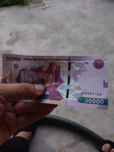 A hand holds a 50,000 Uzbekistani Som banknote. The banknote features architectural imagery and Uzbek text. The background is a textured gray surface, possibly a sidewalk or concrete ground, with a hint of green foliage in the upper left corner. The image appears to be taken outdoors in natural light. There is no indication of the specific location within the city of Mingchinar (Blagoveshchensky), Uzbekistan.