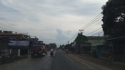 The image depicts a cloudy sky over a street in Blangpeuria, Indonesia. The road appears to be paved, with a dirt shoulder on the right side. Several buildings line the street, including shops and what appears to be a construction site on the left. There are numerous power lines crisscrossing the sky. Several people are visible, most of them riding motorcycles. A red truck is stopped on the left side of the road, and a yellow vehicle, possibly a steamroller, is behind it. Signs with text are visible on some of the buildings. The overall impression is of a busy street in a developing area.