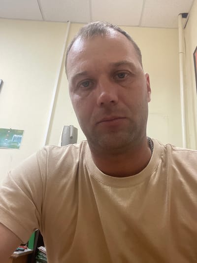 The image shows a close-up, slightly rotated shot of a man's face and upper torso. He has short, graying hair and is wearing a light brown t-shirt with a silver chain around his neck. The man's eyes are open, and he appears to be looking directly at the camera. His expression is neutral, and there are no strong indicators of emotion.
The background is blurry but suggests an indoor setting. To the left, a textured ceiling and a white horizontal beam are visible. The wall behind him is a pale yellow color. On the wall, a small, framed poster with some green and white imagery can be seen. Further to the right, a filing cabinet or shelves with folders are partially visible. A small, gray device is mounted on the wall.
The lighting appears to be artificial, typical of an indoor environment. There are no clear indications of the time of day or weather conditions. The overall scene suggests a casual or possibly office setting. No visible text is legible in the image.