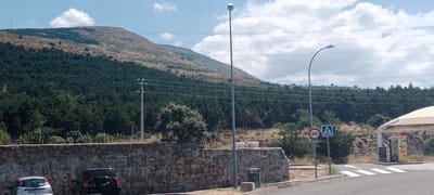The image depicts a rural roadside scene with a mountain covered in trees in the background. A stone wall runs along the foreground, behind which are two parked cars, a light-colored hatchback and a black SUV. To the right of the stone wall, there is a paved area with a crosswalk, a speed limit sign indicating 20, and a pedestrian crossing sign. Further to the right, there is a yellow vending machine or gas pump structure under a light-colored awning. Two tall streetlights are visible, one on the left and one on the right, with power lines stretching across the sky. The sky is partly cloudy. The scene appears to be located in the outskirts of El Espinar, Spain, possibly near a rest stop or service area. There are no people visible in the image.