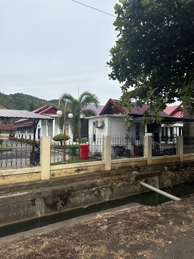 The image captures a scene in the city of Lampisang, Indonesia, focusing on a row of white buildings with red-tiled roofs. A dark metal fence with decorative scrollwork runs along the foreground, separating a paved area from a concrete drainage ditch that contains murky water. Beyond the fence, a cream-colored wall supports the fence. Inside the fenced area, there are several buildings, a palm tree, a decorative lamppost with two spherical lights, a green trash bin, and a red trash bin. A motorcycle is parked near the left side of the image, behind the fence. Overgrown vegetation, including a large tree with broad leaves, partially obscures the view of the buildings and the sky. In the background, lush green hills are visible under a cloudy sky. The drainage ditch in the foreground shows some debris, including a plastic bottle floating in the water.