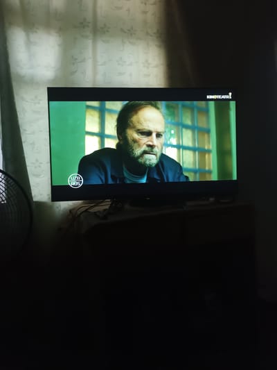 The image shows a television screen displaying a man with a beard, looking intensely. The TV is placed on a dark surface, possibly a table or cabinet, in a dimly lit room. To the left of the TV, a fan is partially visible. The background consists of a curtain with a floral pattern. A logo for "KINO TEATR" and another logo with "UZB RUS" are visible on the screen. The scene is indoors, likely in a residential setting. The city of Kibray, Uzbekistan, is not explicitly depicted in the image.