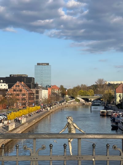 The image is a view of Bydgoszcz, Poland, seen from a bridge over the Brda River. The river runs through the scene, with buildings and a promenade along the left bank. A metal fence with decorative elements and a statue of a male figure leaning over the river are prominent in the foreground. The sky is partly cloudy, with the clouds positioned on the left side of the image. Along the promenade, people are sitting on benches, and trees and shrubs are visible. Buildings of various heights and architectural styles line the cityscape.