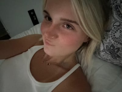 A young Caucasian woman with blonde hair and freckles is lying in bed, looking at the camera. She is wearing a white tank top and a delicate necklace with small beads. The room appears to be dimly lit, with a patterned pillow and a wall-mounted object visible in the background. There is no discernible connection to the city of Squamish, Canada.
