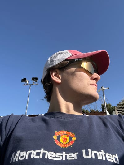 An upward-angled, low-perspective outdoor photograph captures an individual in Hod HaSharon, Israel. The subject, likely male, is visible from the chest upwards, wearing a red-brimmed and white-mesh trucker hat and black-framed 'OAKLEY' mirrored sunglasses that intensely reflect direct sunlight. A navy blue t-shirt with the 'Manchester United' club crest and text is worn on the torso. The individual is looking upwards and to the right. The background features a clear blue sky. On the left, a tall metal pole supports two horn-shaped speakers or floodlights. On the right, another metal pole holds a surveillance camera, with a fence topped by barbed wire positioned below it.