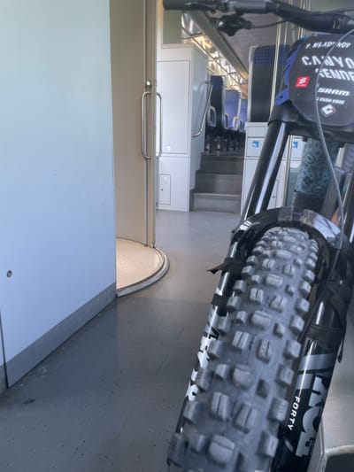 The image depicts the interior of a train carriage, possibly in Merichleri, Bulgaria, though this location is not explicitly visible. A mountain bike is positioned in the foreground, taking up a significant portion of the right side of the frame. The bike's front wheel and suspension fork are prominently featured, with the "FOX 40" branding visible on the fork. The background shows rows of empty blue and gray train seats, suggesting the train is either at a station or during a quiet period of travel. The lighting is artificial, typical of an indoor train environment. To the left of the bike, a white wall dominates the space, with a partially open door revealing a curved walkway and a glimpse of the train's exterior or another section of the interior. The floor is a dark, speckled material. There are no people clearly visible in the image. The overall impression is one of travel and possibly the anticipation of an outdoor activity, given the presence of the mountain bike.