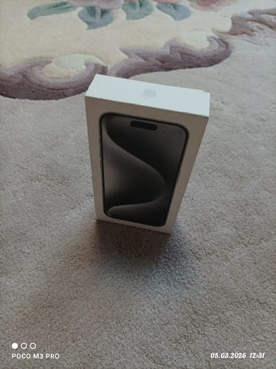 The scene is located indoors in Dmitrievskaya, Russia, captured on March 5, 2026, at 12:31, by a POCO M3 PRO device. A white rectangular retail box, identified by an Apple logo on its visible top surface, stands upright on a light beige, textured carpet. The front of the box displays an image of a dark smartphone featuring a dynamic island and a wavy abstract wallpaper. The carpet exhibits an ornate floral and scrollwork pattern in shades of purple and green towards the upper background. A distinct shadow is cast by the box onto the carpet towards the lower right. No individuals are present, and no actions or interactions are depicted within the static scene.