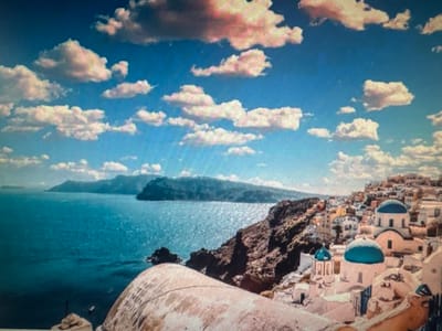 This media file depicts a picturesque coastal scene with a village nestled on a hillside overlooking the sea. The foreground features a large, textured architectural element, possibly a dome or a rounded building, with a rough, light-colored surface. Beyond this, a vibrant blue sea stretches out towards a distant, hazy coastline with rolling hills.
The village itself is a prominent feature, characterized by whitewashed buildings with distinctive blue-domed roofs. The architecture is clustered tightly on the steep incline, creating a charming and iconic Mediterranean or Aegean aesthetic. Small pathways and steps wind through the village.
The sky above is a brilliant blue, adorned with fluffy white clouds, some tinged with pink and orange, suggesting either early morning or late afternoon light. The sun appears to be bright, casting a warm glow on the scene and creating highlights on the water's surface. The sea is calm, with gentle ripples reflecting the sunlight. The overall mood is tranquil and idyllic, conveying a sense of peace and beauty.
While no people are clearly visible, the presence of boats on the water and the inhabited nature of the village suggest human activity. The location, with its characteristic architecture and coastal setting, strongly resembles Santorini, Greece, a popular tourist destination known for its white villages and blue domes. There is no visible text in the image. The image does not contain any elements that specifically relate to Ellwangen, Germany.
