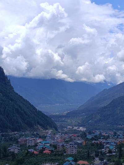 This is a vertical, eye-level, outdoor shot of a valley in Palchan, India, under a cloudy sky. The valley is filled with a dense village that spreads out below steep, tree-covered mountains. The mountains on the left are dark green with pine trees, while the mountains in the background are a muted blue, suggesting distance and elevation. The sky is filled with dramatic, bright white and gray clouds, with patches of blue sky visible. The village consists of numerous multi-story buildings with a variety of roof colors, including red, teal, and gray. The buildings are clustered together, with roads winding through them. The foreground features more dense greenery and some partially constructed buildings. The overall impression is one of a vibrant, inhabited valley nestled within a grand mountainous landscape.