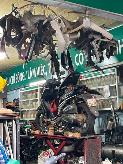The image depicts the interior of a motorcycle repair shop, likely located in Liên Nghĩa, Vietnam. The scene is cluttered and functional, filled with motorcycle parts and equipment. In the foreground, a black and red scooter is elevated on a red hydraulic lift. Various plastic fairings and metal components are hanging from the ceiling above, creating a dense canopy. Shelves filled with bottles of engine oil, possibly Castrol, are visible in the background, along with other tools and machinery. A green banner with white Vietnamese text, which appears to be a motto or slogan, is stretched across the wall. The overall impression is one of a busy workshop where repairs and maintenance are actively carried out. There are no people visible in the image.