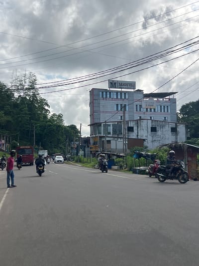 The scene depicts a street in Paragoda, Sri Lanka, lined with parked motorcycles and moving traffic. The view is slightly angled. A tall, weathered building with the sign "M. MAWELI Group of Company" is visible. Power lines crisscross the sky above. A group of people is present at the bottom of the frame, with one individual in a pink shirt standing next to a motorcycle. A red three-wheeled vehicle (Tuk-Tuk) and other cars are seen on the road. Lush green trees line parts of the road. The sky is overcast with gray clouds.