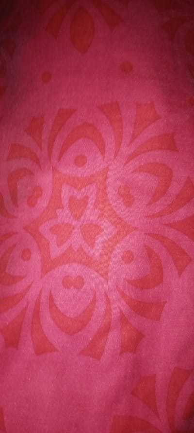 A close-up shows a red fabric with an ornate pattern in a slightly lighter shade of red. The design incorporates curved shapes, dots, and elements resembling stylized flowers or leaves. The fabric texture has a subtle sheen. The patterns are organized in a radial arrangement, creating a sense of repeating motifs across the material.
