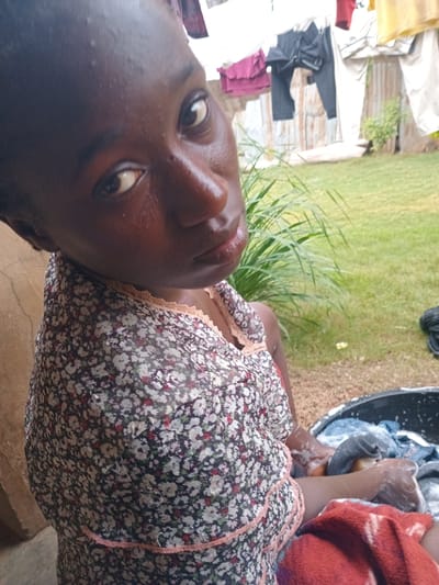 This media file shows a close-up, rotated shot of a young Black person in an outdoor setting. The person's face fills much of the frame, with their eyes and nose clearly visible. They appear to be looking upwards and towards the viewer. Their skin is dark and appears slightly wet, with small beads of moisture visible. They are wearing a floral-patterned top with a lace trim around the neckline.
In the background, to the right of the person, is a black basin filled with soapy water and laundry. Hands, also appearing wet and covered in soap suds, are actively engaged in washing clothes within the basin. A red and white patterned cloth is draped over the person's shoulder and arm.
To the left of the person, out of focus, are some laundry items hanging on what appears to be a clothesline or fence. The ground appears to be made of concrete or a similar hard surface, and there is a patch of green grass in the background. The overall lighting suggests it is daytime, likely sunny, as indicated by the shine on the person's skin. The scene suggests a domestic chore, specifically laundry being done outdoors. The location context provided is Bokkos, Nigeria. No text is visible in the media file.