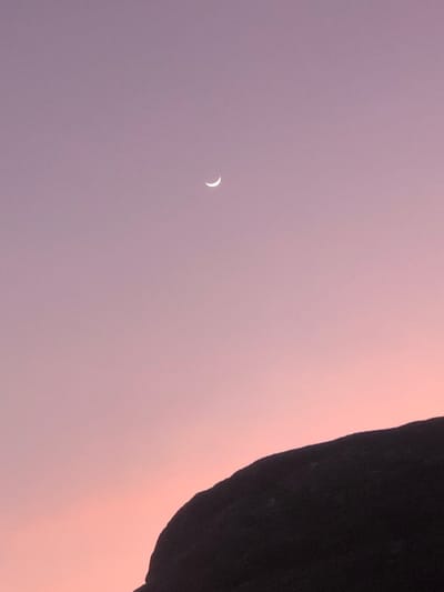 A dusky pink and purple sky is the backdrop for a crescent moon. The moon is small and positioned on the left side of the frame. A dark, rugged object, likely a rock formation, occupies the upper right corner. This object is a dark, almost black color, contrasting with the soft hues of the sky. The overall composition creates a sense of serene isolation.