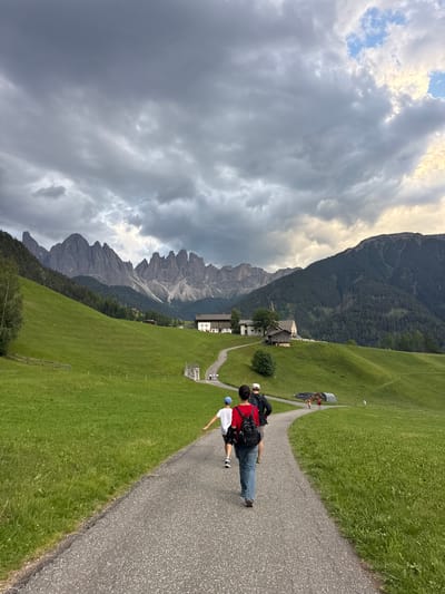 The image shows a scenic view of the Italian Alps, likely near the village of St. Magdalena (Santa Maddalena), Italy, known for its picturesque church steeple and surrounding Dolomite peaks. The foreground features a paved pathway curving through lush green rolling hills. Several people are walking on the path, appearing to be on a hike or stroll. In the middle ground, there are scattered buildings, possibly farmhouses or guesthouses, nestled into the landscape. The background is dominated by the dramatic, jagged peaks of the Dolomites, with a cloudy sky overhead that suggests approaching or receding weather. The overall impression is one of natural beauty and outdoor activity in a mountainous region.