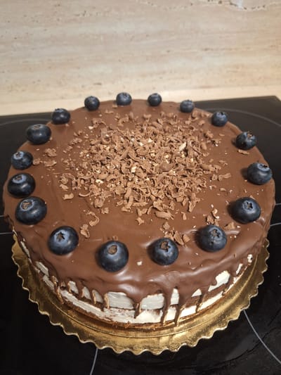 This is a close-up, overhead shot of a chocolate cake topped with blueberries and chocolate shavings. The cake is on a golden, scalloped cake board and appears to be a layered cake with white frosting between the chocolate cake layers. Chocolate ganache is drizzled over the top and sides of the cake, with some drips extending down the sides. The cake is positioned on a dark, reflective surface, likely a stovetop, and a beige, marbled countertop is visible to the left. The scene is indoors and well-lit, suggesting it could be a kitchen setting. No specific time of day, weather, or emotions are discernible. There is no visible text in the image. The cake is the sole subject of the photograph.