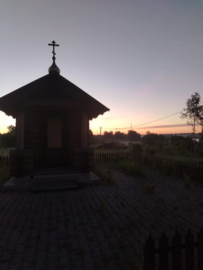 The image, captured in Petrovskoye, Russia, presents an outdoor scene at either sunrise or sunset, characterized by a low-light silhouette effect. In the left foreground, a small, dark wooden chapel with a steep, gabled roof stands, its structure appearing as a dark mass against the sky. A golden onion dome, surmounted by a dark Orthodox cross, crowns the chapel's roof. The chapel rests on a multi-tiered base of lighter-toned stone or concrete blocks. The immediate ground in front of and to the right of the chapel is paved with dark, interlocking hexagonal or square pavers, with sporadic patches of green grass growing between them. A low, dark wooden picket fence extends across the mid-ground, separating the paved area from an expanse of dense, overgrown vegetation including bushes and trees. A thin power line stretches horizontally across the upper mid-ground, and distant utility poles are faintly visible against the horizon. The background features a soft, colorful sky transitioning from light grayish-blue at the top to hues of pink, orange, and pale yellow near the horizon, with a subtle layer of mist or fog visible over the distant landscape. A distinct, lighter-barked tree with dark branches is present on the far right. No individuals are depicted in the scene.