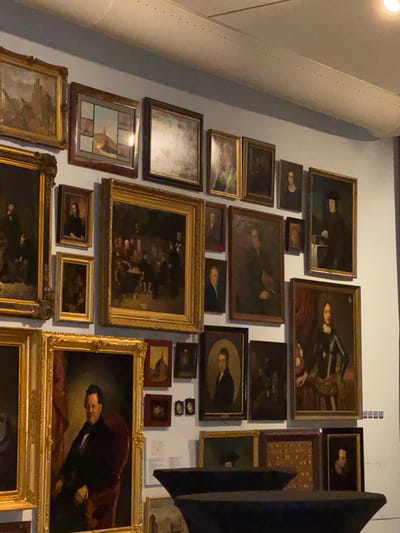 This image displays a wall covered with numerous framed paintings, suggesting an art gallery or museum setting. The paintings vary in size and frame style, with many featuring dark, ornate gold frames. The subjects of the paintings are predominantly portraits of individuals, often depicted in formal attire. Some paintings appear to be historical or religious in nature, with figures in period clothing and rich, dark backgrounds. One prominent painting in the lower right features a man in a dark suit and a flowing red cloak, his gaze directed downwards and slightly to the side, conveying a somber or introspective mood. Other paintings show smaller groups of people or abstract scenes. The lighting in the image suggests an indoor environment, likely artificially lit, with some reflections visible on the glass of the frames. The overall impression is one of a curated collection of art, densely displayed on a plain wall, with a large, possibly structural, cylindrical element running up the left side of the frame. There is no visible text within the image itself.