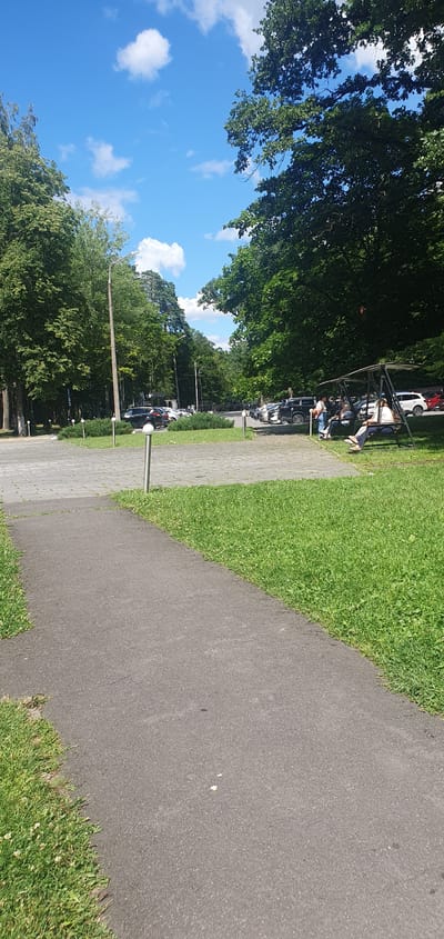 A paved pathway leads diagonally through a grassy area towards a parking lot filled with cars. In the background, a line of trees forms a dense canopy. To the right of the pathway, on the grass, a swing set with a canopy is occupied by four individuals. The sky is blue with scattered white clouds. The overall scene suggests a park or recreational area within a suburban or semi-rural environment, possibly in the vicinity of Чонкаўскі сельскі Савет, Belarus.