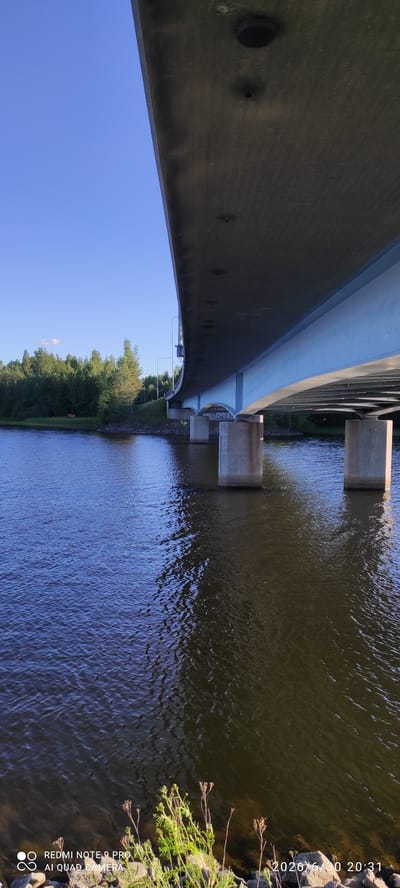 This media file depicts a view from underneath a concrete bridge looking out over a body of water. The scene is oriented vertically, with the sky at the bottom and the water at the top.
The underside of the bridge is a prominent subject, showing its concrete structure with some circular indentations and what appear to be metal fixtures. The bridge curves upwards and away from the viewer. Support pillars for the bridge are visible in the water, with concrete structures extending upwards from them.
The water is dark and shows ripples, suggesting a gentle movement. On the right side of the image, the rocky bank of the waterway is visible, with some dry vegetation and bright green plants growing. On the left side, a dense line of green trees creates a distinct treeline along the opposite bank of the water. The sky is a clear, bright blue.
The image appears to be taken in the late afternoon or early evening, as suggested by the timestamp and the quality of light. The weather seems to be fair and sunny.
Visible text includes a timestamp and camera information: "2026/6/30 20:31" and "REDMI NOTE 9 PRO AI QUAD CAMERA". The location is identified as Hartola, Finland.
There are no people or animals visible in the frame. The overall mood is serene and natural, focusing on the architecture of the bridge and the surrounding environment.