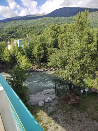 The image captures a scenic view of a mountain landscape with a river flowing through it. In the foreground, a turquoise railing of a bridge or elevated walkway dominates the left side, casting shadows from a sunny sky. Beyond the railing, a fast-flowing river, appearing somewhat turbulent, cuts through a rocky bank.
The river is bordered by lush green trees on both sides, with dense foliage covering the landscape. Behind the trees, a large industrial building with a light-colored facade and a blue section is visible, suggesting a town or industrial area nestled within the valley. Tall chimneys rise from the building.
Further in the background, a majestic mountain range covered in thick green forest rises towards the sky. The sky itself is a mix of blue with scattered white clouds, indicating pleasant weather. The sunlight suggests it is daytime, likely late morning or early afternoon.
On the right bank of the river, near the trees, there are two signs. The sign on the right appears to have the word "DANGER" visible in red letters at the top, with other text below, though it is not fully legible. Next to the signs, a pile of dry brushwood lies on the grassy ground. The overall scene conveys a sense of natural beauty interspersed with human industrial presence. The location is identified as Saint-Michel-de-Maurienne, France.