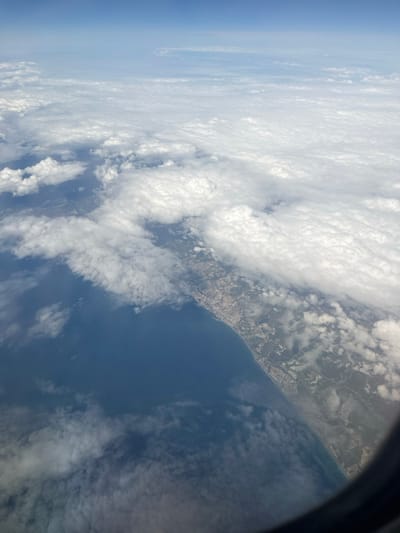 An aerial photograph, captured from an aircraft window, presents a high-altitude view of a coastal region under a partly cloudy sky. The deep blue ocean occupies the left and lower-center portions of the image, meeting a distinct, curving coastline that extends from the upper-right to the lower-center-right. Along this coastline, a continuous strip of urban development, characterized by numerous structures and infrastructure, is visible. Inland from the urbanized zone, the terrain appears as a mix of lighter-toned land and some green patches, partially obscured by cloud cover.
Extensive cloud formations dominate the atmosphere, ranging from fluffy white cumulus clouds scattered over the landscape to more expansive, flatter white and grayish cloud layers that obscure significant parts of the land and extend towards the horizon in the upper and right sections of the frame. Above these clouds, a clear, light blue sky transitions to a deeper blue at the top edge of the image. The dark, curved outline of an aircraft window frame is visible in the bottom right corner, partially vignetting the scene.
No people or discernible actions are present. The visible geography, featuring a prominent coastline with substantial urban development, does not correspond to the inland location of Nzalat Laadam, Morocco.