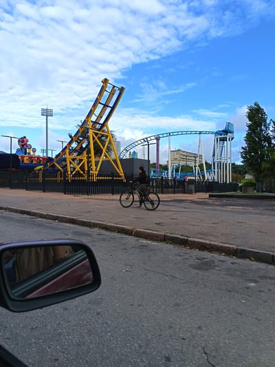 The image shows a partial view of a street in Montevideo, Uruguay, with amusement park rides in the background. A woman is riding a bicycle on the street. In the foreground, the side mirror of a car captures a partial reflection of the interior. The amusement park rides include a yellow and blue roller coaster, a blue roller coaster, and a tower with stadium lighting in the background. The sky is blue with scattered clouds.