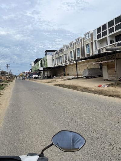 The image depicts a street scene in the city of Landuh, Indonesia. The foreground shows the handlebar and rearview mirror of a motorcycle, indicating the perspective of the photographer. The road ahead is paved and appears to be a main thoroughfare, with a dusty shoulder on the left side.
Along the right side of the road, a row of multi-story buildings with modern architecture lines the street. These buildings feature a mix of white, brown, and green facades, with decorative elements on the upper floors. Some of the ground-floor storefronts are closed with metal shutters. A small wooden kiosk with a sign that reads "Pos. Laundry" is visible in front of one of the buildings. A car covered with a gray tarp is parked in front of another building. Several motorcycles are parked along the sidewalk.
On the left side of the road, there are more modest buildings and what appears to be a less developed area with some vegetation and utility poles with numerous overhead wires. A few vehicles, including motorcycles and a dark-colored car, are present on the road, moving in both directions. The sky is overcast with light clouds. The overall impression is a developing urban environment in Indonesia.