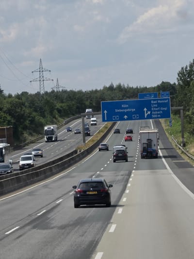 This is a full, eye-level shot of a multi-lane highway with several vehicles moving in both directions. The highway is flanked by trees on the left and right, with a concrete barrier separating the lanes. Power lines and transmission towers are visible in the background. The sky is overcast with some clouds.

Specifically, this image captures a section of the A3 Autobahn near Windhagen, Germany. A large blue overhead sign indicates directions to Köln (Cologne) and Siebengebirge straight ahead, and to Bad Honnef, Linz, Eitorf (Sieg), and Asbach 500m to the right. Several cars and a large white truck are visible on the highway. The license plate of the black Peugeot in the foreground is visible and reads "KK 332-B". The road surface shows signs of wear and some darker patches, possibly from recent rain.