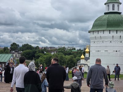The image depicts a gathering of people outdoors, with a large white building with a green domed roof dominating the right side of the frame. This building appears to be a historical or religious structure, possibly a church or monastery, given the presence of golden domes in the background. In the distance, a cityscape with trees and buildings can be seen under a cloudy sky.

Several individuals are visible in the foreground and mid-ground, appearing to be tourists or visitors. They are of various ages and are dressed in casual clothing. Some are walking, while others are standing and observing the surroundings. One woman in the foreground is wearing a headscarf, common attire for women visiting religious sites in some cultures.

The atmosphere seems somewhat subdued, possibly due to the overcast weather. The sky is filled with heavy, grey clouds, suggesting a chance of rain or a generally cool, overcast day. The presence of many people suggests an event or a popular tourist destination.

The text "КОФЕ С СОБОЙ" is visible on a sign attached to a small building on the left side of the image, which translates to "COFFEE TO GO." This indicates a point of sale for beverages within the complex.

The overall scene suggests a visit to a cultural or historical site in Sergiyev Posad, Russia, on a cloudy day. The interaction between the people and the architecture suggests a typical tourist experience.