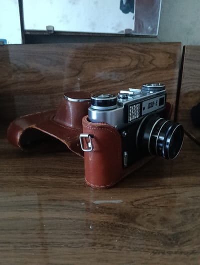 A vintage Zenit-5 camera with its original leather case is placed on a wooden surface. The camera is silver and black, with a prominent lens and a metal textured panel above the lens. The leather case is a reddish-brown color and has a vintage appearance. The wooden surface appears to be a table or shelf with a visible wood grain pattern. The background is out of focus, but suggests an indoor environment with a reflective surface, possibly a mirror or window, in the upper portion of the image. The scene is lit by artificial light, casting subtle shadows. No people are present. The overall impression is one of a still life, showcasing a classic piece of photographic equipment. The specific location in the city of Florești, Moldova cannot be determined from the image alone.