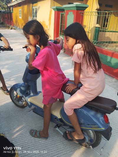 Two young girls interact with a light blue/grey electric scooter on a concrete pavement in Wanguk, Indonesia. The girl on the left, wearing a pink t-shirt and shorts, stands straddling the scooter, her right hand on the left handlebar grip, and her left elbow resting on the handlebars, with her chin in her hand, looking downwards. She wears light grey sandals. The girl on the right, wearing a light pink short-sleeved jumpsuit, sits on the scooter's brown rear seat, also looking downwards and slightly to the right. She wears black flip-flops and a multi-colored beaded bracelet on her left wrist. A third person's arm and hand are visible on the far left, holding handlebars of another vehicle, out of frame. The scooter features a cream-colored base seat, a red taillight, and visible text "ADEE SAVE ENERGY 2.0" on its side panel. In the background, a yellow single-story building with white-framed windows stands on the left. To the right of that, another building features red-painted structural elements and a light green metal fence. The scene is illuminated by bright daylight, casting distinct shadows on the ground and buildings. The ground is a light grey concrete surface with horizontal grooves.