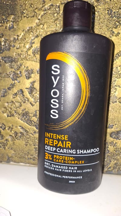 A black plastic bottle of Syoss Intense Repair Deep Caring Shampoo is positioned vertically on a white surface. The bottle has a dark brown cap and a label with white and orange text. The text on the label reads "SYOSS EST. OSAKA, JAPAN 1977", "INTENSE REPAIR DEEP CARING SHAMPOO", "3% PROTEIN-CARE-COMPLEX", "DRY, DAMAGED HAIR REPAIRS HAIR FIBERS IN ALL LEVELS", and "PROFESSIONAL PERFORMANCE". Below this, a number "2999334" is visible. The background is a textured, mottled golden-yellow wall. There are no people or animals present in the image. The location is not specified as Voinova, Moldova.