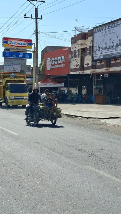 In Keudebayu, Indonesia, a man wearing a dark hooded sweatshirt drives a motorcycle with a trailer attached. The trailer is laden with green coconuts and carries three young boys. The boy in the middle wears a black and white soccer jersey with "Jeep" written on it. They are traveling on a paved road under a bright blue sky with visible power lines overhead. To the left, a yellow truck is parked. In the background, a row of buildings displays various signs, including "Indomaret," "GODA SAFER. SMART. EV," and "UNITED BIKE." A tent with the "YADEA MOTOR LISTRIK PINTAR" logo is also visible. The environment appears to be a commercial or roadside area.