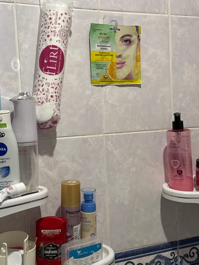 The image shows a collection of personal care products on a tiled wall in a bathroom. On the top left, a large pack of "Flirt" cotton pads is hanging from a hook. To its right, a single-use face mask packet labeled "Viteks Vitamin Active" is also hanging. Below these, on a white shelf, are various bottles and tubes of toiletries, including a white dispenser, a bottle of "NIVEA" body lotion, and a tube of hand cream. On a lower shelf, several more items are visible: a gold-capped spray bottle labeled "Рексона," a blue-capped spray bottle labeled "soft cotton," a red deodorant stick with "Old Spice" branding, and a small clear plastic container with what appear to be dental floss picks. To the right, on another shelf, is a pink bottle of "STORIE D'AMORE" body wash. The tiled wall and the arrangement of products suggest a domestic bathroom setting. The location is specified as Яромінскі сельскі Савет, Belarus.