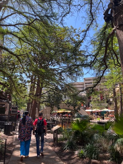 This image shows a section of the San Antonio River Walk in Texas, USA. In the foreground, two pedestrians walk along the paved walkway. One person wears a colorful patterned shirt and blue pants, while the other wears a red shirt and jeans, with a backpack. The walkway is bordered by metal railings and planters with vegetation. Beyond the walkway, there is a canal area, likely part of the San Antonio River, with outdoor dining areas visible. Tables with umbrellas and people seated are scattered along the waterfront. Buildings are visible in the background. Overhead, trees with green foliage provide shade. A lighting rig is secured in one of the trees.