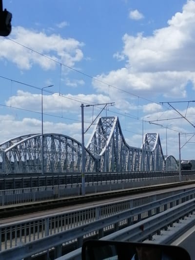 The image depicts a large, metal truss bridge spanning across a body of water, with railway tracks visible on the bridge. The sky above is blue with scattered white clouds. The bridge's structure is characterized by its intricate network of beams and supports, painted in a light color, likely gray or white. Overhead, electrical wires for trains are strung between poles. In the foreground, a road or highway with guardrails is visible, suggesting a perspective from a vehicle traveling alongside or on an approach to the bridge. In the distance, on the bridge, a vehicle can be seen. The scene appears to be set on a clear day, with good visibility. Based on the architectural style of the bridge and its infrastructure, this image likely portrays a section of the Mureș River Bridge in Stelnica, Romania, which carries both road and rail traffic. There are no identifiable individuals in the visible portion of the image.