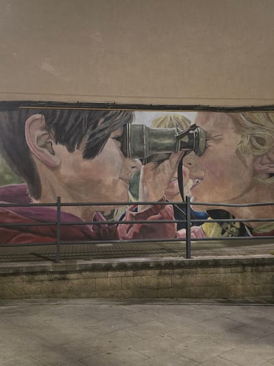 The image displays an outdoor mural on a building wall in Cornellà De Llobregat, Spain. The mural, painted in a realistic style, depicts three young individuals in profile, primarily focusing on two figures interacting with a telescope or spyglass. The leftmost figure, with short dark hair, wears a maroon top and looks into the eyepiece of an antique-style, possibly brass or oxidized green, telescope. The figure on the far right, with blonde, wavy hair, looks into the opposite end of the telescope, with their mouth slightly open. A third partial face is visible between these two, positioned behind the telescope.
A dark grey metal railing spans horizontally across the lower portion of the mural, partially obscuring the artwork. Below the railing, a low wall constructed from light brown, rectangular-cut stone blocks forms a base. In the foreground, a grey paved sidewalk, composed of square tiles, is visible, showing scattered small debris. Above the mural, the building wall is a plain light brown, with multiple bundles of dark electrical wires and a thin yellow conduit running horizontally across it. Shadows from the railing and unseen overhead elements are cast onto the stone wall and sidewalk, indicating artificial light sources in a low-light environment.