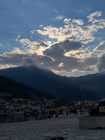 The image depicts a view in Skala Potamias, Greece, facing the mountains and harbor. The foreground consists of a paved stone wall. Beyond, there's a view of the harbor filled with boats. Houses and buildings line the harbor. Tall mountains rise in the background, partly obscured by clouds. The sky is blue with fluffy, light-colored clouds. Light streams through gaps in the clouds, illuminating the mountains. A lamppost stands beside the harbor.